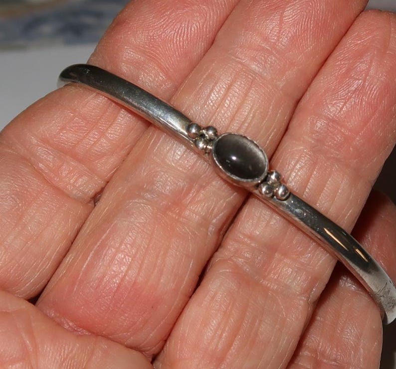 May include: A silver-toned metal bangle bracelet featuring a smooth, polished band. The center of the bracelet is adorned with a single oval-shaped dark gray stone, flanked on both sides by three small, decorative silver-toned metal spheres.