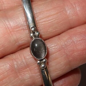 May include: A silver cuff bracelet featuring a smooth, polished metal band. The center of the bracelet is adorned with a single oval-shaped, dark gray stone set in a bezel. Small, round silver spheres are clustered on both sides of the stone.
