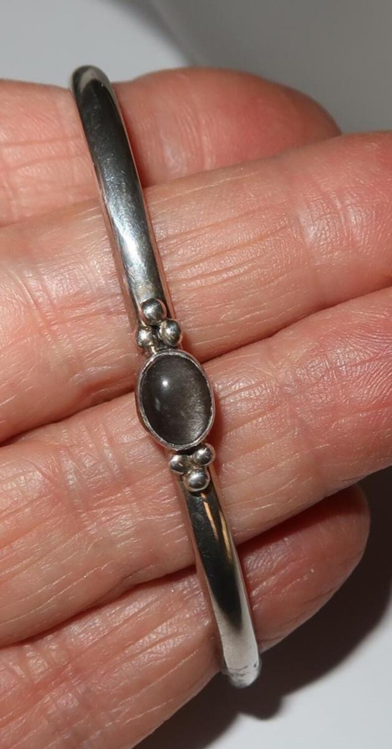 May include: A silver cuff bracelet featuring a smooth, polished metal band. The center of the bracelet is adorned with a single oval-shaped, dark gray stone set in a bezel. Small, round silver spheres are clustered on both sides of the stone.