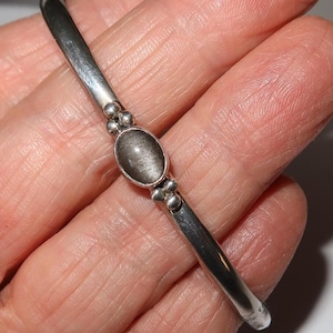 May include: A silver-toned metal bangle bracelet featuring a central oval-shaped gray stone with a chatoyant sheen. The stone is flanked by small decorative metal spheres on both sides, set into a smooth, polished, and rounded metal band.
