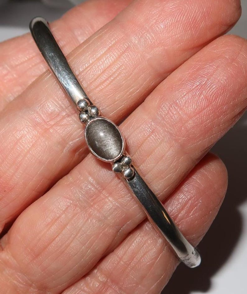 May include: A silver-toned metal bangle bracelet featuring a central oval-shaped gray stone with a chatoyant sheen. The stone is flanked by small decorative metal spheres on both sides, set into a smooth, polished, and rounded metal band.
