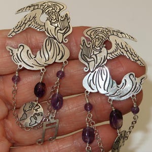 May include: A pair of silver-toned angel earrings. Each earring features an angel playing a harp, with dangling amethyst beads, a musical note charm, and a small globe charm. The earrings are detailed with etched designs.