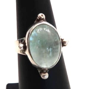 May include: A silver ring with an oval, light green gemstone. The ring has a simple band and is accented with small, round silver details on the sides and top.
