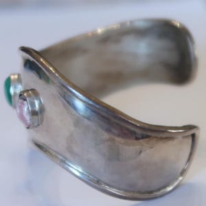 May include: A wide, polished silver-toned cuff bracelet featuring a smooth, curved band. Two circular bezel-set stones are mounted on the side, one being a solid green cabochon and the other a pale pink, marbled stone with a smooth, rounded surface.