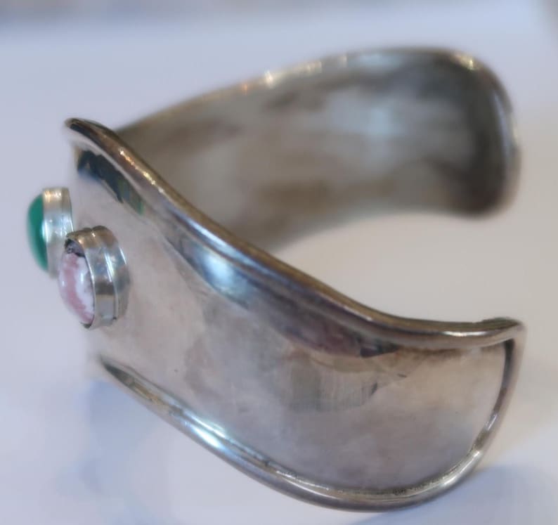 May include: A wide, polished silver-toned cuff bracelet featuring a smooth, curved band. Two circular bezel-set stones are mounted on the side, one being a solid green cabochon and the other a pale pink, marbled stone with a smooth, rounded surface.