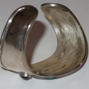 May include: A wide, open-ended silver cuff bracelet with a brushed, textured interior surface. The exterior features a smooth, polished finish with a dark, rounded stone accent visible on one side and a small turquoise stone on the other.