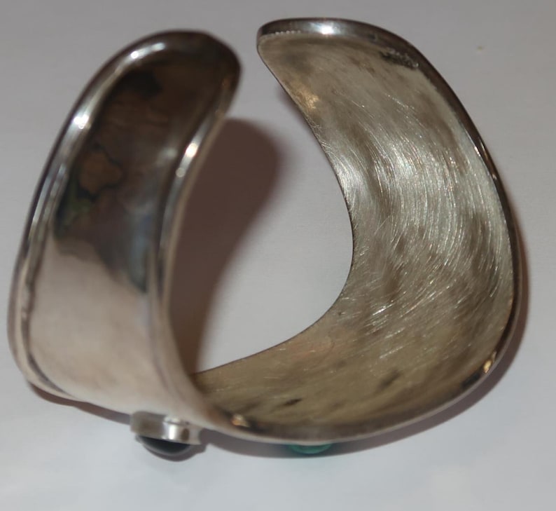 May include: A wide, open-ended silver cuff bracelet with a brushed, textured interior surface. The exterior features a smooth, polished finish with a dark, rounded stone accent visible on one side and a small turquoise stone on the other.
