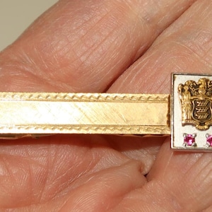 May include: Gold-toned tie bar with a rectangular design. The bar features a textured pattern and a rectangular emblem with a coat of arms and two small, square, red gemstones. A classic accessory.