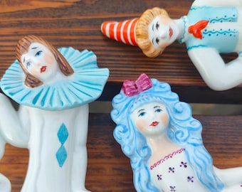 May include: Three porcelain figurines of people with painted details. One figurine is a woman with blue hair and a white dress with a floral pattern. Another figurine is a woman with brown hair and a blue and white costume. The third figurine is a person with blonde hair wearing a blue and white costume and a red and white hat.