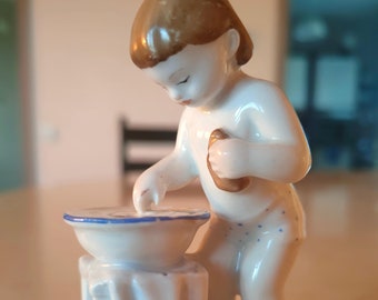 May include: A porcelain figurine of a child holding a small bowl, standing on a pedestal with a white cloth draped over it. The figurine is painted in white and blue.