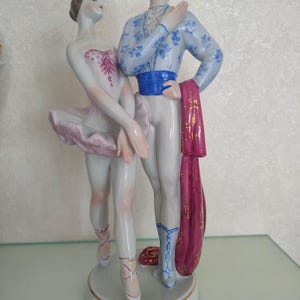 May include: A porcelain figurine of two ballet dancers standing on a circular base. The dancer on the left wears a white leotard with pink accents and a pink tutu. The dancer on the right wears a blue patterned top, blue sash, and white tights.