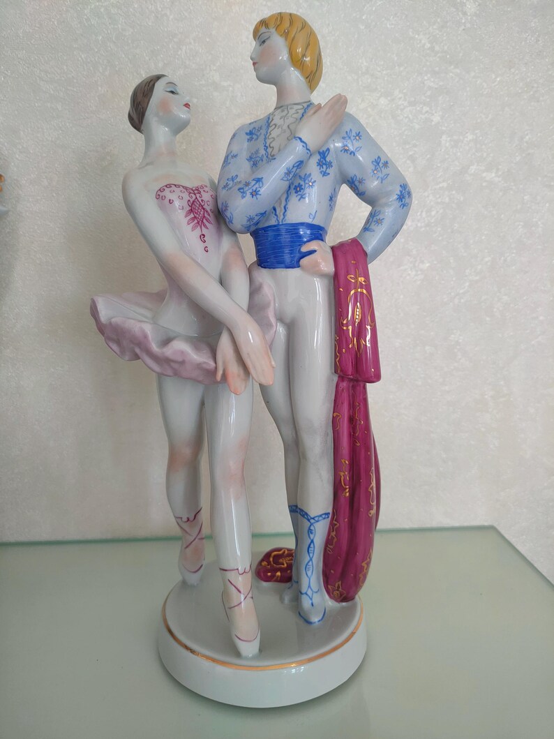 May include: A porcelain figurine of two ballet dancers standing on a circular base. The dancer on the left wears a white leotard with pink accents and a pink tutu. The dancer on the right wears a blue patterned top, blue sash, and white tights.