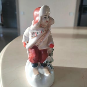 Pode incluir: Uma figurinha de porcelana de uma pessoa vestindo um chapéu vermelho e branco e um casaco branco com detalhes dourados. A pessoa está em pé em esquis e tem um cachecol verde e branco. A figurinha está em uma base branca.