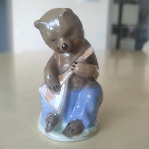 Pode incluir: Uma estatueta de cerâmica de um urso marrom tocando uma balalaica. O urso está usando calças azuis e está sentado. A balalaica é branca com detalhes dourados e laranja. A estatueta está em uma base branca.