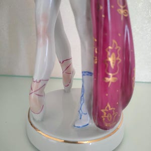 May include: A porcelain figurine showing the lower legs of two dancers standing on a circular base. One dancer wears white ballet slippers with pink laces, while the other wears a blue patterned boot. A deep magenta fabric with gold floral details hangs.