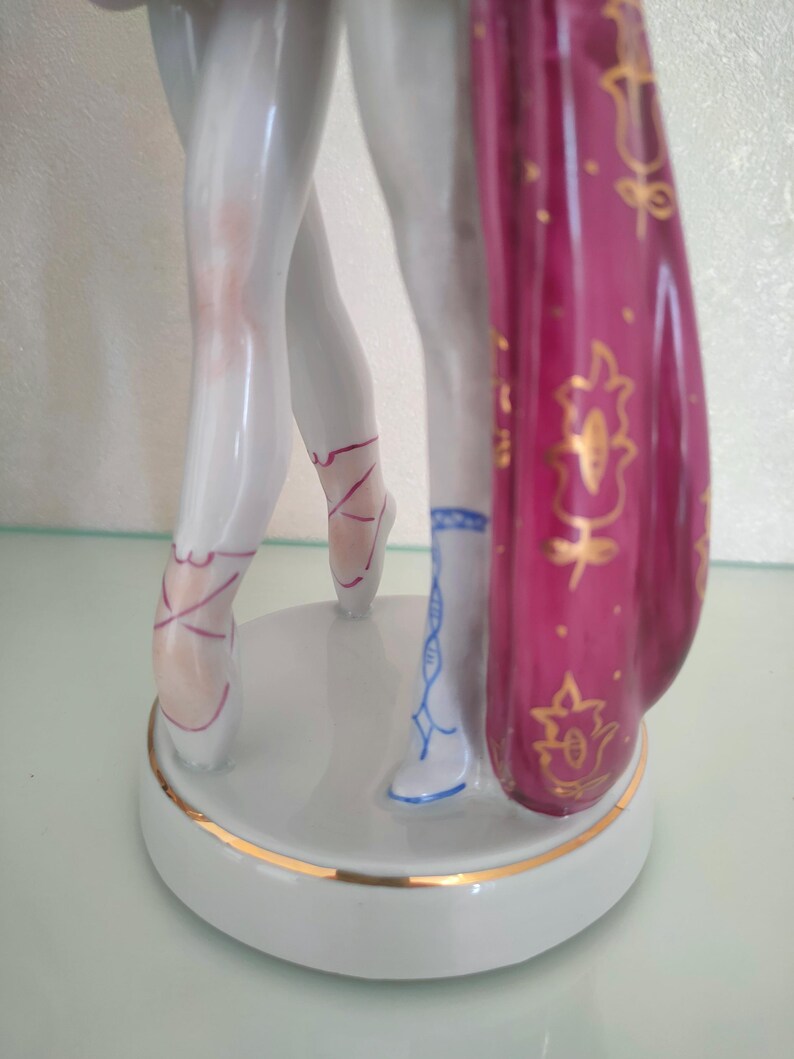 May include: A porcelain figurine showing the lower legs of two dancers standing on a circular base. One dancer wears white ballet slippers with pink laces, while the other wears a blue patterned boot. A deep magenta fabric with gold floral details hangs.