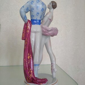 May include: A porcelain figurine of two ballet dancers in a pose. The male dancer wears a light blue top with blue floral patterns and a bright blue sash. The female dancer wears a pink tutu. A long magenta fabric drape with gold patterns hangs down.