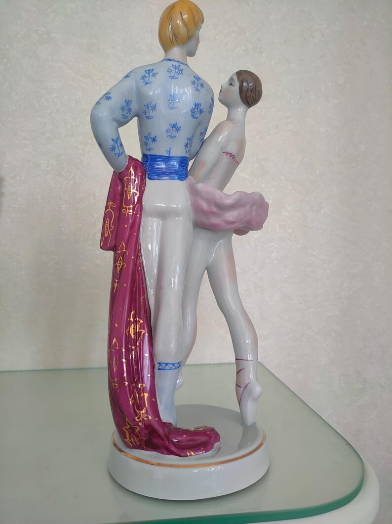 May include: A porcelain figurine of two ballet dancers in a pose. The male dancer wears a light blue top with blue floral patterns and a bright blue sash. The female dancer wears a pink tutu. A long magenta fabric drape with gold patterns hangs down.
