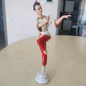 Może przedstawiać: Porcelanowa figurka tancerki w tradycyjnym stroju. Figurka jest w kolorze białym, czerwonym i złotym. Tancerka jest w ruchu, z jedną nogą wyciągniętą i ramionami wyciągniętymi. Podstawa jest biała ze złotymi wykończeniami.