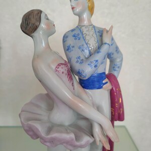 May include: A porcelain figurine of two dancers in a pose. The female figure wears a pale pink tutu and bodice with red detailing. The male figure has blonde hair and wears a light blue patterned jacket, a blue sash, and holds a dark pink cloth.