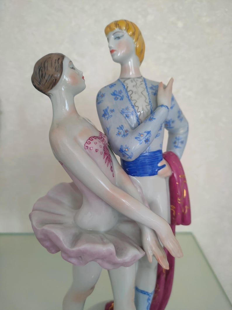 May include: A porcelain figurine of two dancers in a pose. The female figure wears a pale pink tutu and bodice with red detailing. The male figure has blonde hair and wears a light blue patterned jacket, a blue sash, and holds a dark pink cloth.