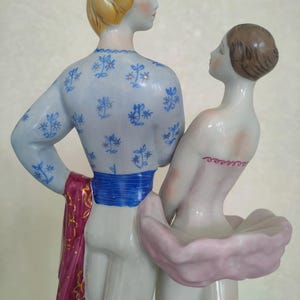 May include: A porcelain figurine of two figures standing together. The taller figure has blonde hair and wears a light blue top with a blue floral pattern, a bright blue sash, and a dark pink cloth. The shorter figure has brown hair and wears a pink tutu.