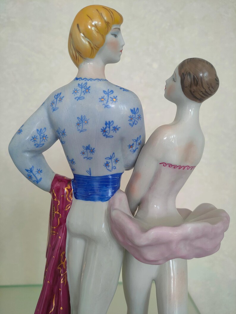 May include: A porcelain figurine of two figures standing together. The taller figure has blonde hair and wears a light blue top with a blue floral pattern, a bright blue sash, and a dark pink cloth. The shorter figure has brown hair and wears a pink tutu.