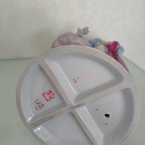 May include: The underside of a porcelain figurine base featuring a white glazed finish with a cross-shaped support structure. It displays the red printed number 37, a red stylized letter K logo, and the red text C 23P, along with a small circular hole.