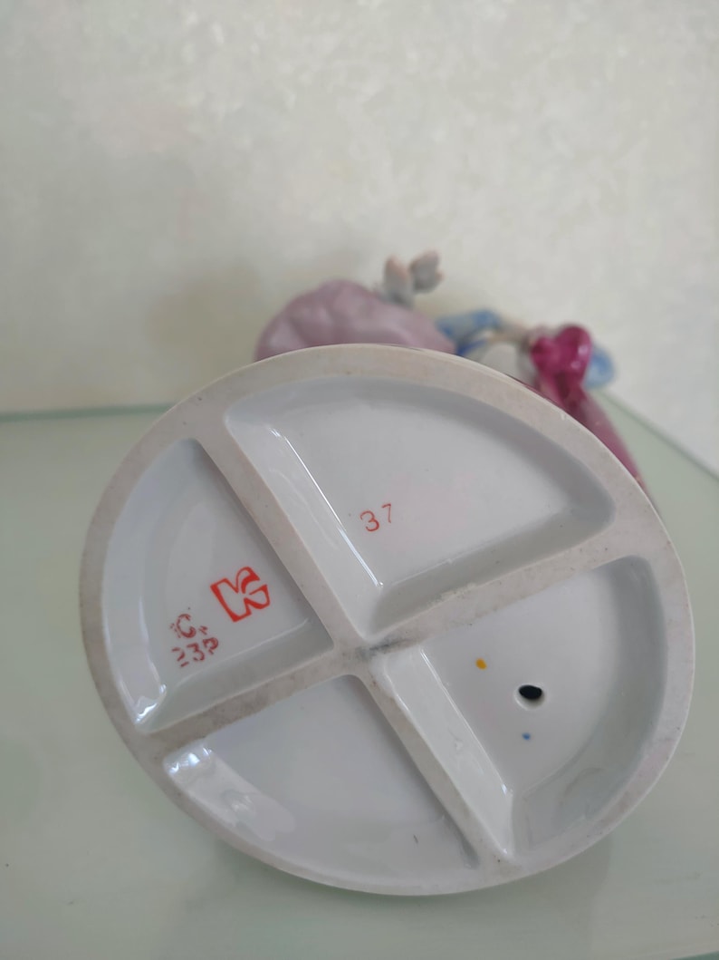 May include: The underside of a porcelain figurine base featuring a white glazed finish with a cross-shaped support structure. It displays the red printed number 37, a red stylized letter K logo, and the red text C 23P, along with a small circular hole.