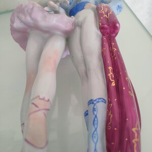 May include: A porcelain figurine depicting two figures standing together. One figure wears a pale pink tutu and ballet slippers with pink laces. The other figure wears blue patterned trousers, a blue sash, and holds a long, deep magenta cape.