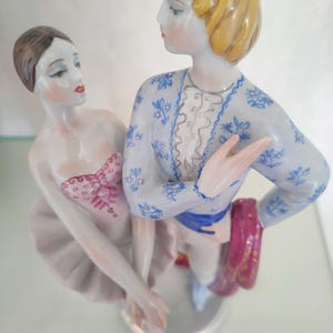 May include: A porcelain figurine of two ballet dancers standing together. The female dancer wears a pale pink tutu and bodice with red detailing. The male dancer has blonde hair and wears a light blue jacket with a blue floral pattern and a red sash.