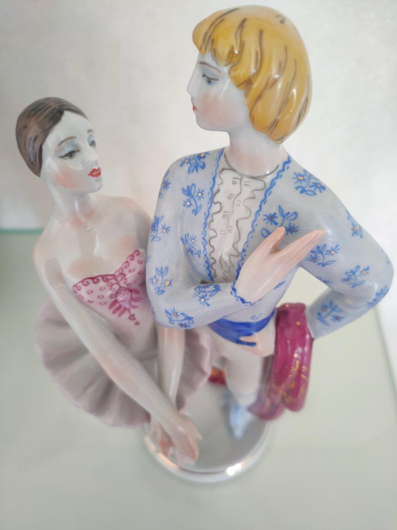 May include: A porcelain figurine of two ballet dancers standing together. The female dancer wears a pale pink tutu and bodice with red detailing. The male dancer has blonde hair and wears a light blue jacket with a blue floral pattern and a red sash.