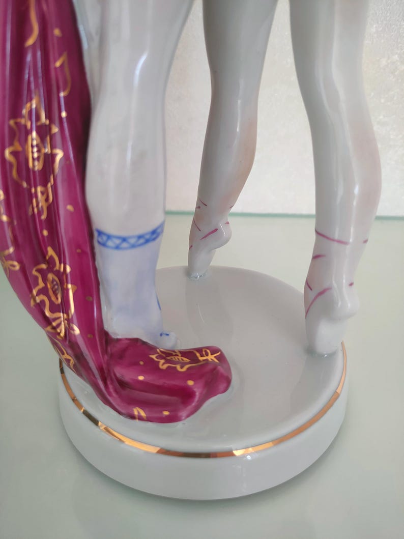 May include: A porcelain figurine showing the lower legs of two dancers standing on a circular white base with a gold rim. One figure wears a deep magenta fabric with gold floral patterns, while the other has light pink ballet slipper ribbons.