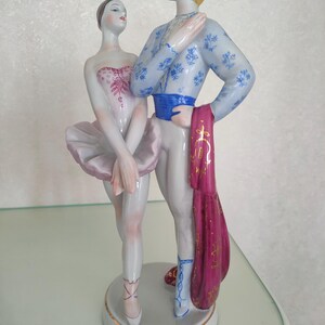 May include: A porcelain figurine of two ballet dancers standing on a circular base. The female dancer wears a pale pink tutu and bodice, while the male dancer wears a light blue floral patterned top, a blue sash, and holds a magenta cloth.