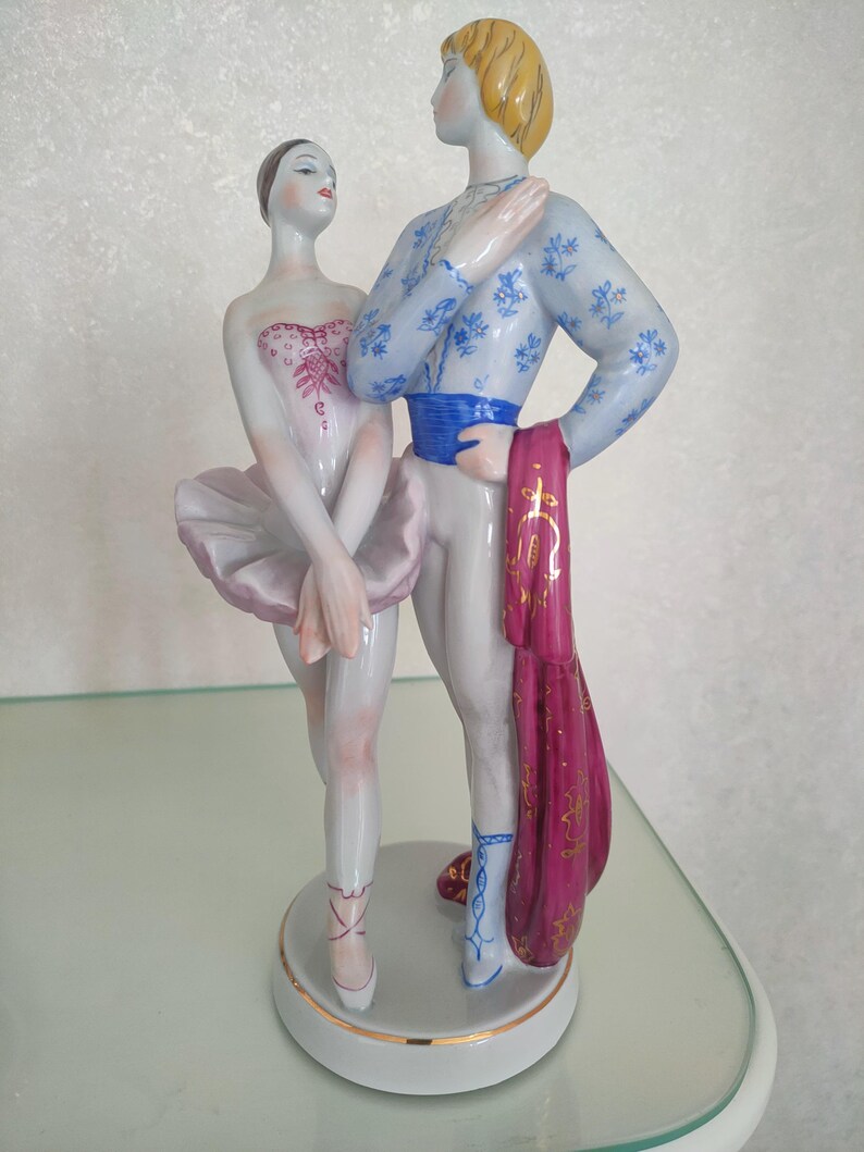 May include: A porcelain figurine of two ballet dancers standing on a circular base. The female dancer wears a pale pink tutu and bodice, while the male dancer wears a light blue floral patterned top, a blue sash, and holds a magenta cloth.