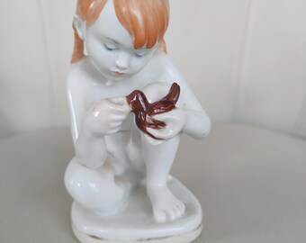 May include: A white porcelain figurine of a young person with auburn hair, gazing down at a brown bird held in their hands. The figurine is in a seated position, with a smooth, glossy finish.