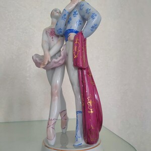 May include: A porcelain figurine of a ballet dancing couple standing on a circular base. The dancer wears a pale pink tutu and bodice, while the partner wears a light blue patterned jacket, white trousers, and a long magenta cape with gold trim.