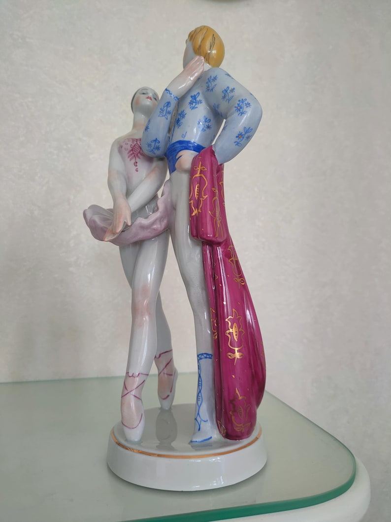 May include: A porcelain figurine of a ballet dancing couple standing on a circular base. The dancer wears a pale pink tutu and bodice, while the partner wears a light blue patterned jacket, white trousers, and a long magenta cape with gold trim.