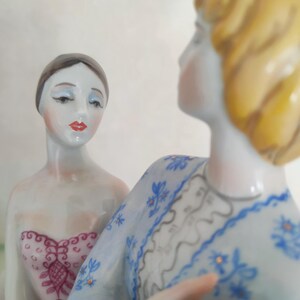 May include: Two porcelain figurines featuring a figure with dark hair in a pink patterned bodice and a figure with blonde hair wearing a light blue long-sleeved garment with a blue floral pattern and a white lace-like collar detail.