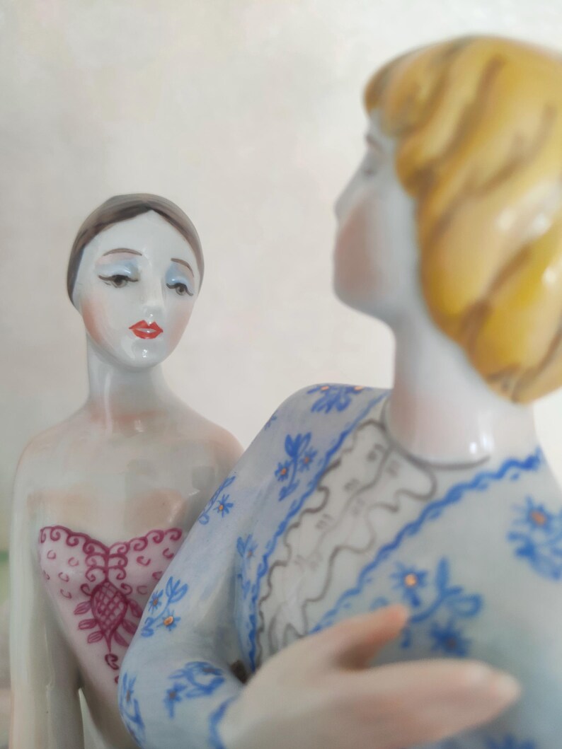 May include: Two porcelain figurines featuring a figure with dark hair in a pink patterned bodice and a figure with blonde hair wearing a light blue long-sleeved garment with a blue floral pattern and a white lace-like collar detail.