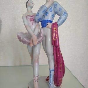 May include: A porcelain figurine depicting a ballet dancer and a partner standing together. The dancer wears a pale pink tutu and bodice, while the partner wears a light blue patterned top, a blue sash, and holds a long, deep magenta fabric drape.
