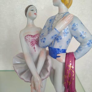 May include: A porcelain figurine of two ballet dancers standing together. The dancer on the left wears a white tutu and a pink bodice with intricate detailing. The dancer on the right wears a light blue patterned top, a blue sash, and a magenta cloth.