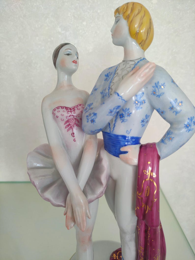 May include: A porcelain figurine of two ballet dancers standing together. The dancer on the left wears a white tutu and a pink bodice with intricate detailing. The dancer on the right wears a light blue patterned top, a blue sash, and a magenta cloth.