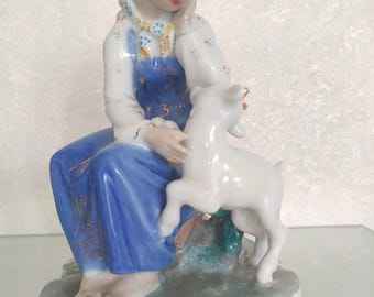 May include: A porcelain figurine of a seated figure wearing a white blouse with a yellow patterned scarf and a blue skirt with gold accents, resting a hand on a small white goat. The figure has a blue headband and sits on a gray base.