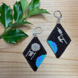 May include: A pair of diamond-shaped beaded earrings featuring a black background with pixelated designs. One earring displays a silver circular object and a blue crescent, while the other shows a silver lightning bolt and a blue crescent shape.