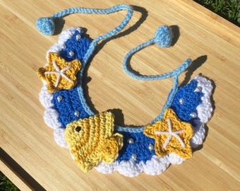 May include: A crocheted necklace with a blue and white wave pattern, yellow fish and starfish accents, and white pearl beads.