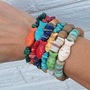 May include: A collection of colorful beaded bracelets. The bracelets feature various stone beads and fish-shaped charms in shades of orange, turquoise, red, green, and yellow. The bracelets are stacked on a wrist.