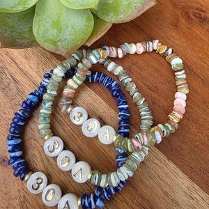 May include: Three beaded bracelets with the text "30A" in gold lettering. The bracelets feature a mix of colorful beads in shades of blue, green, pink, and white. The bracelets are displayed on a wooden surface.