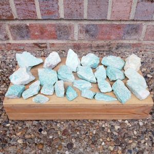 May include: A collection of raw, unpolished amazonite stones in varying shades of pale blue, teal, and white. The stones are angular, rough-textured, and display natural crystalline structures, arranged together on a rectangular wooden block.