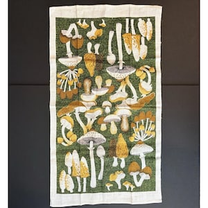 May include: A rectangular kitchen towel featuring a repeating botanical print of various mushrooms in shades of brown, tan, and white against a dark green background. The fabric has a visible weave texture and a light-colored border on all sides.